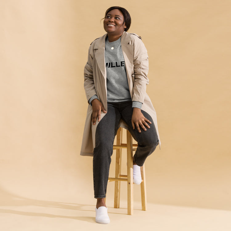 Sudadera MILLE Origin LS de color gris carbón con logo MILLE en el pecho. Vista de cuerpo entero con modelo sonriendo y sentada en un taburete.