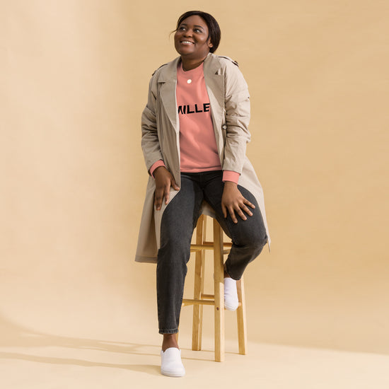 Sudadera MILLE Origin LS de color rosa empolvado con logo MILLE en el pecho. Vista de cuerpo entero con modelo sonriendo y sentada en un taburete.